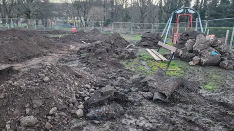 Mark Mather Lots of earth is piled up with big boulders next to a dirty blue and red swing. The playpark looks like a construction site and is surrounded by metal fencing