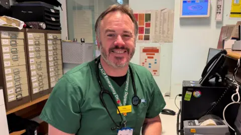Somerset NHS Foundation Trust A male paramedic with grey hair and a grey beard is wearing dark green scrubs and sitting at a reception desk smiling at the camera.