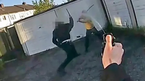A still from police bodycam footage of a police officer holding up a baton as he confronts a man holding a long curved sword and wearing a yellow hooded top. The arm of the officer who is wearing the camera can be seen spraying pepper spray