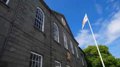 Photo of the Guernsey States Chamber building. It's a grey granite block building with with white-paned windows. The sky is blue and there is a Guernsey flag on a pole in front of the building flapping in the wind.