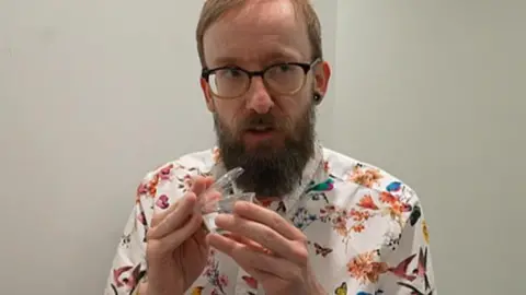 A man with glasses and a beard holding a plastic pot in his hands. He is wearing a watch and a shirt which has birds and flowers on.