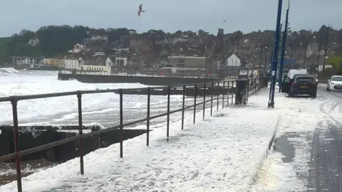 Robin Red Breast A pavement and road running next to a beach, with foamy water pouring from the sea side out onto the road.