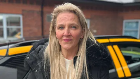 Adele Cook, a 55-year-old white woman with long blonde hair wearing a black padded coat. She is standing in front of an East Midlands Ambulance Service vehicle