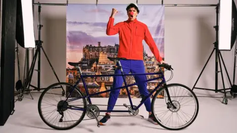 PA Media Radio 1 DJ Greg James stood behind a black tandem bike with his left hand on the handlebars and his right arm in the arm with his fish clenched. He is wearing blue trousers and a red jacket and has a red nose on his nose, with a white cap. The photo is taken in a studio setting so there are two big bright photography lights behind him and a canvas style picture of Edinburgh Castle.