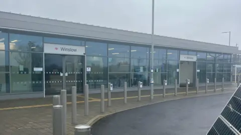 Diana Blamires An empty train station which has two signs with "Winslow" on them. There is a row of posts in front of the entrance which is mainly glass fronted. There is a solar panel on the right hand side of the picture.