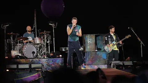 Getty zdjęcia trzech członków Coldplay na scenie w USA. Chris Martin jest w środku z perkusistą Will Will po prawej i basistę Guy Berryma po jego lewej stronie.