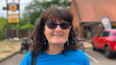 Chrissie Alexander is standing outdoors in a car park. She is looking directly at the camera and smiling and is wearing a pair of glasses over her eyes and a blue T-shirt with a bag over her right shoulder.
