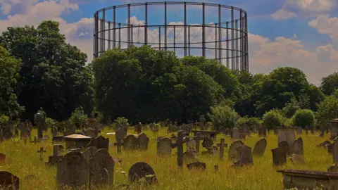 Getty Images Kensal Green Cemetery with graves hidden in overgrown grass and flowers. In the background are trees with a large gasholder in the skyline.