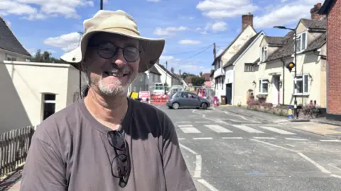 Matt Marvel/BBC A man stands beside a village road on a sunny day. He is smiling and wears a large cream hat and a grey T-shirt. Sunglasses rest around the rim of the T-shirt and on his chest. He has glasses on and a white beard. The road behind him has been closed off due to roadworks.