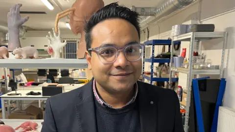 Nazmus Tareque from Anthrotek. He is standing inside an industrial unit where there are rows and tables with prosthetic items including silicon rats, body parts and organs. He is looking directly at the camera and smiling and is wearing a red and blue shirt with a blue jumper and dark blazer over the top.
