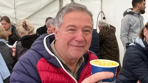 The photo shows supporter Mark Duffy smiling, with a cup of spicy butternut squash soup, wearing a navy and red puffer jacket.