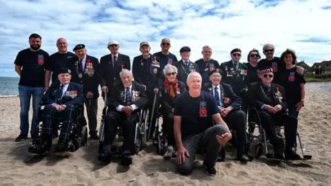 Peter Smoothy 18 people on a beach. There are five elderly people in wheelchairs, 13 people stood behind them. One man kneeling at the front