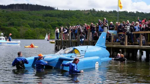 PA Media Crowds gather on a wooden jetty and take photographs as Bluebird floats on the water next to them, attended by crew members.