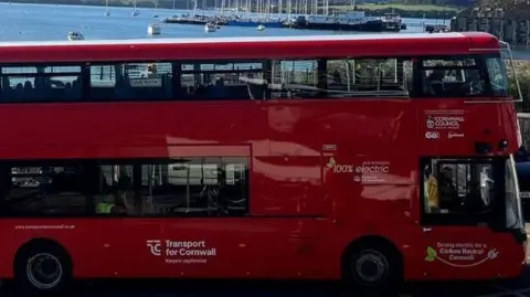 Cornwall Council A red double decker bus in Cornwall. The vehicle is electric. A harbour with several boats on the water is in the background.