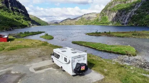 Karen Davies Image of a motorhome which has been parked alongside a large mountain lake. In the background can be seen a small red hut and a lake surrounded by steep hills and mountains. 