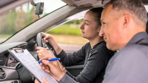 A young woman sitting her driving test. A male instructor is marking her on a clipboard