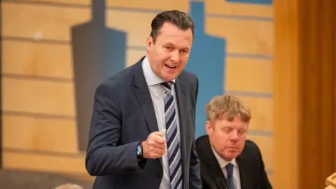 PA Media Russell Findlay stands in the Holyrood debating chamber and speaks and gestures. He has shot dark hair and gestures 