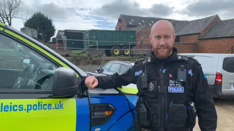 A policeman stands in uniform, with his hand on his police 4x4 vehicle. In the background you can see farm buildings and farm machinery.