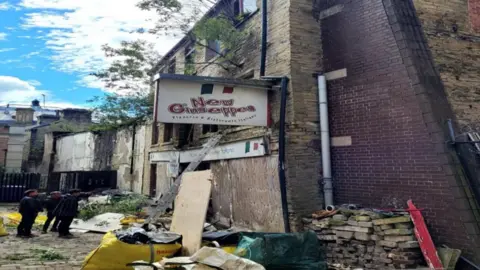 Three men stand outside a crumbling stone building with a sign on the wall that reads 'New Giuseppes'