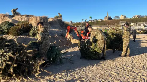 Members of RAF Lossiemouth in camouflage clothing moving Christmas trees across the sand on a sunny day
