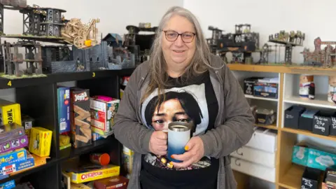 A woman with long grey hair and glasses is wearing a grey hoodie and holding a blue mug. She is standing in front of shelves containing board games and gaming models.