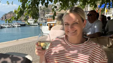 Rachel Bevan Rachel Bevan pictured holding a glass of white wine in her left hand. Rachel wears a red and white striped t-shirt, has blonde hair pulled back into ponytail, blue eyes and smiles with her teeth. A harbour can be seen behind her on the left. Tables filled with people at a restaurant can be seen behind her, sitting below trees in the shade. 