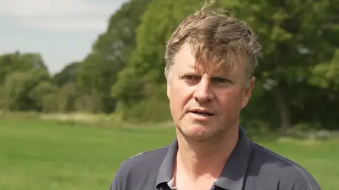 A man with short blonde hair wearing a grey polo shirt. He is stood in a green field with green trees behind him. He is looking off to the left of the camera