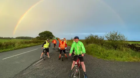 Open Country A group of people on tandem bikes, wearing reflective cycling gear and helmets pose by the side of a road, next to a field, under a rainbow in the sky.