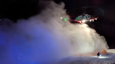 A helicopter preparing to land on Glencoe. The helicopter is white and red and is hovering in mid-air. A large cloud of snow has been kicked up below it. In the foreground, a person is kneeling with a torchlight.