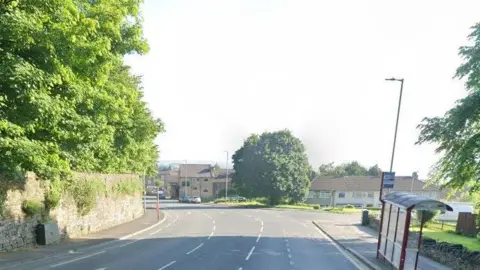 Google A wide suburban road on a sunny day. Leafy trees can be seen, along with a bus stop, a street light, and in the far distance, some houses and cars.