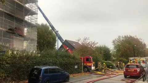 A fire engine with a long ladder next to the tower block, which is covered in scaffolding. A number of firefighters are visible in the picture. 