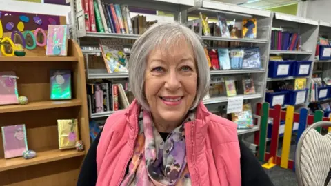 Ollie Conopo/BBC Woman wearing a pink top standing in front of shelves of books