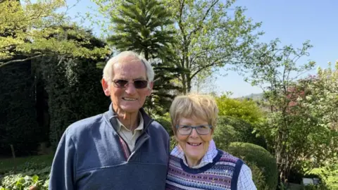 An elderly couple smiling at the camera are standing in a garden, with the Wollemi pine behind them among a range of other trees. The sun is shining and there is also a blue sky in the background.