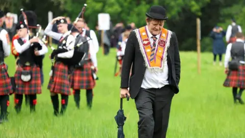 Reuters A man is walking in a field with an umbrella in his right hand. He is looking down at the ground, wearing a dark suit, black bowler hat and an orange sash around his shoulders.