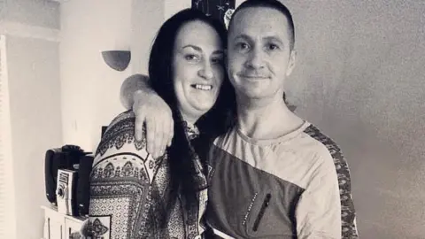 A black and white photo of a couple, David and his wife, smiling and hugging. In casual clothes at home