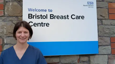 Dr Rebecca Geach is pictured standing in front of a sign for Bristol Breast Care Centre. She's wearing hospital scrubs and has short brown hair.