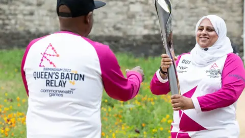 PA Media Rougie Khanom (right) handing the Queen"s Baton to Batonbearer Steven Oram at the Tower of London in London, England