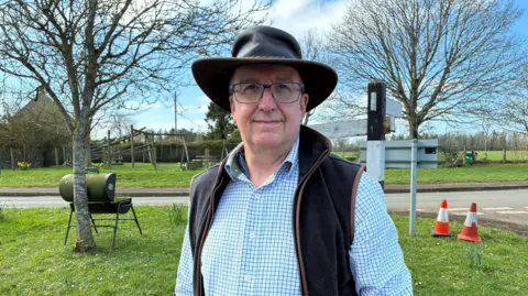 Roger Baskerville stands by a play park. He is wearing a checked shirt and brown gilet and hat. There is a road behind him and several trees.
