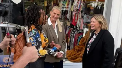 Supplied Three women are being filmed standing outside a charity shop. The woman on the left has black dreadlocks tied in a bun, a vibrant shirt of gold, white and black and black trousers. The woman in the middle has long blonde hair, a white shirt and sage blazer. The woman to the right is actor Joanna Page and has short blonde hair, a thick black coat and a flowery top. Someone out of shot is holding a large camera with two hands. Behind them is a large glass front to a shop filled with racks of clothes.