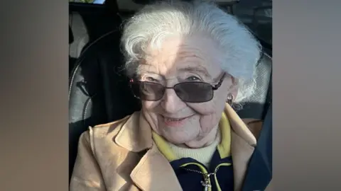 South Yorkshire Police/Family handout A head-and-shoulders image of an elderly woman with grey hair smiling at the camera, peering over sunglasses. She is wearing a camel coloured coat over a navy top with a yellow collar and a cream jumper underneath. She wears a cross on a chain around her neck.
