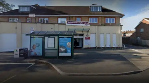 Google Langley Library street view. There is a bus stop in front of the building. Above the entrance, a sign reads "Welcome to Langley Library". It's a sunny day.