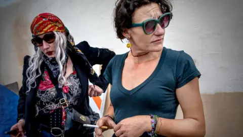 Getty Images Two women who described using drugs, including fentanyl. One wears sunglasses and a blue t-shirt and is holding a cigarette. The other wears sunglasses, a long black coat, and a red bandana.