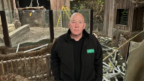 Tony Binskin in a black Wingham Wildlife Park, stood in front of an empty zoo enclosure. Lots of damaged pipework can be seen on the floor.