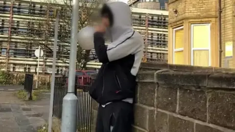 BBC/Charles Heslett A young person wearing a hooded top and tracksuit bottoms, standing against a wall on a street corner and inhaling from a balloon. Their face is pixelated.