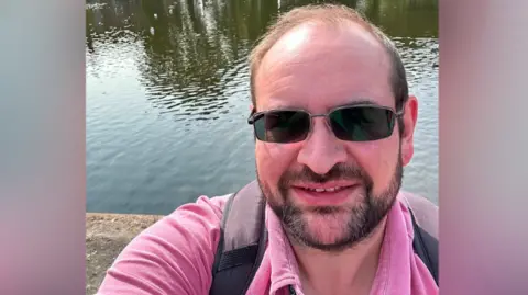 Iain Wallis A head shot of a man with brown hair, a dark beard, dark sunglasses and a pink polo shirt standing in front of the crammer when it was full of water.