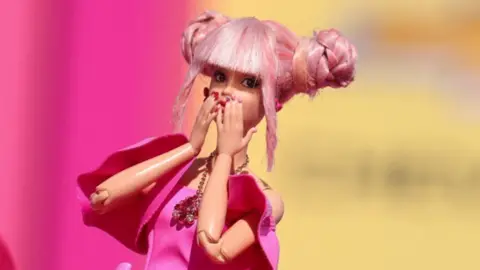 A Barbie doll wearing a pink dress with huge bows on the hips blowing kisses on a red carpet style background
