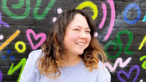 Amber Amber, with dark curly hair, smiles as she leans against a black wall covered in multi-coloured symbols drawn in graffiti style