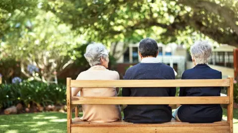 Getty Images Three older people, seen from behind, sat side-by-side on a bench on a sunny day
