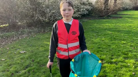 Ollie Conopo/BBC Boy wearing hi-viz vest with "Isaac's Litterpick Group" written on it, He is standing in a park with a litter picker.