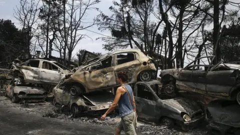 Getty Images A woman walks past the burnt cars after wildfires hit the village of Mati near Athens, Greece on July 24, 2018